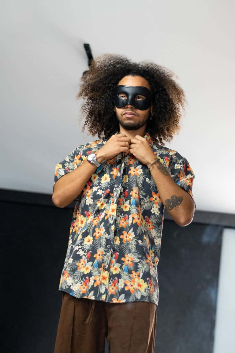 Imagem de um homem com máscara, cabelo afro e camisa floral, em ensaio fotográfico artístico.