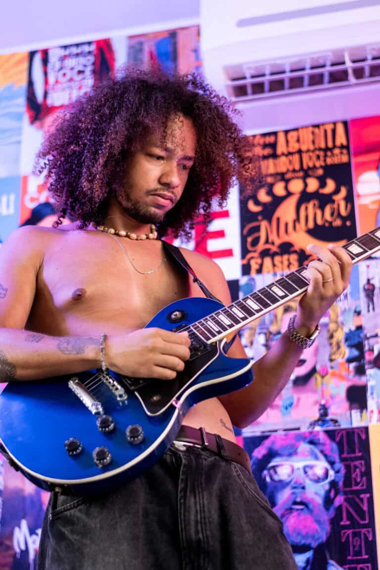 Guitarrista tocando guitarra elétrica em show ao vivo, com fundo de pôsteres coloridos.