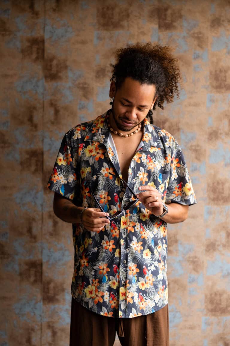 Homem com camisa floral e cabelo cacheado, segurando óculos, em estúdio com fundo texturizado.