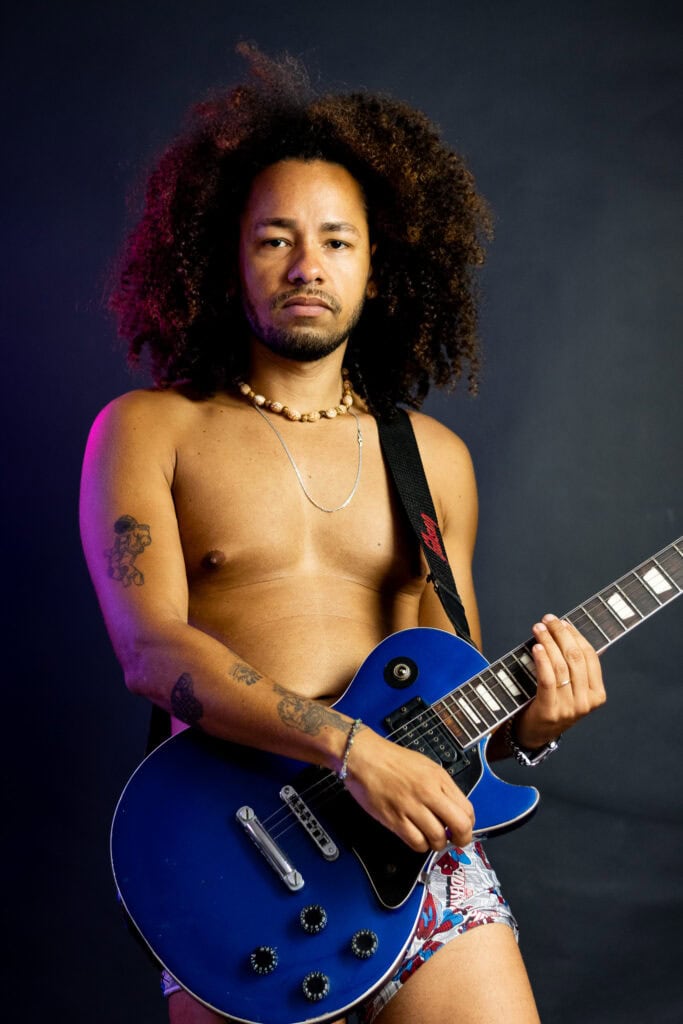 Guitarrista masculino com cabelo afro, sem camisa, segurando guitarra azul, em estúdio profissional.
