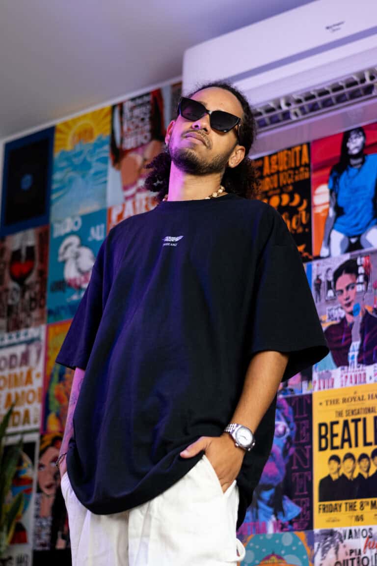 Imagem de um homem com óculos escuros, camiseta preta e calça branca, em ambiente com pôsteres color.