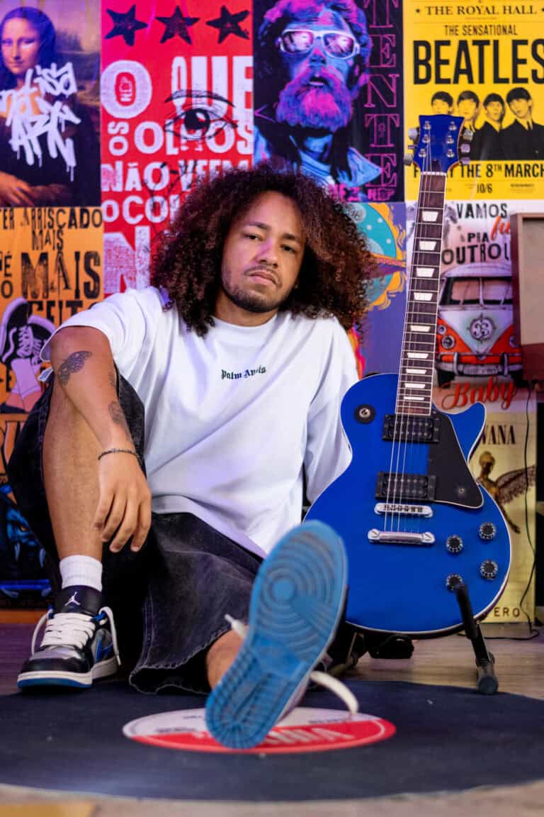 Retrato de um músico com guitarra azul, em ambiente de estúdio com pôsteres de bandas clássicas ao f.