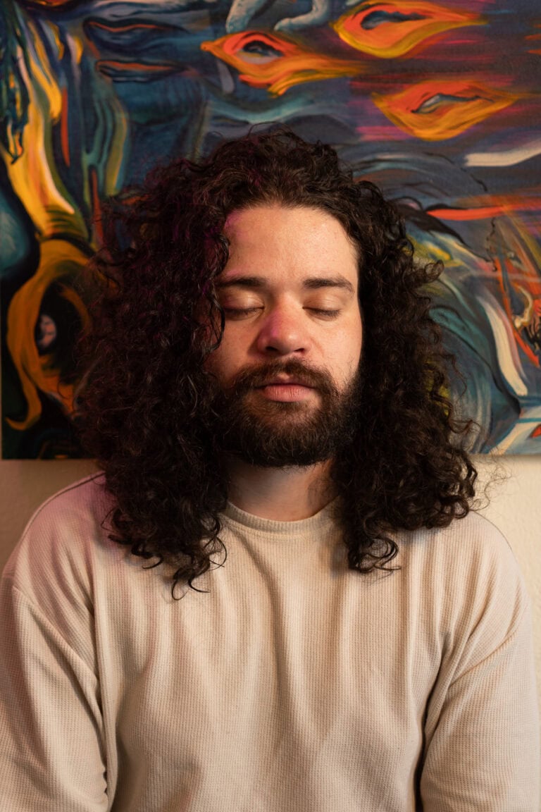 Homem com cabelo cacheado e barba, fundo artístico colorido.