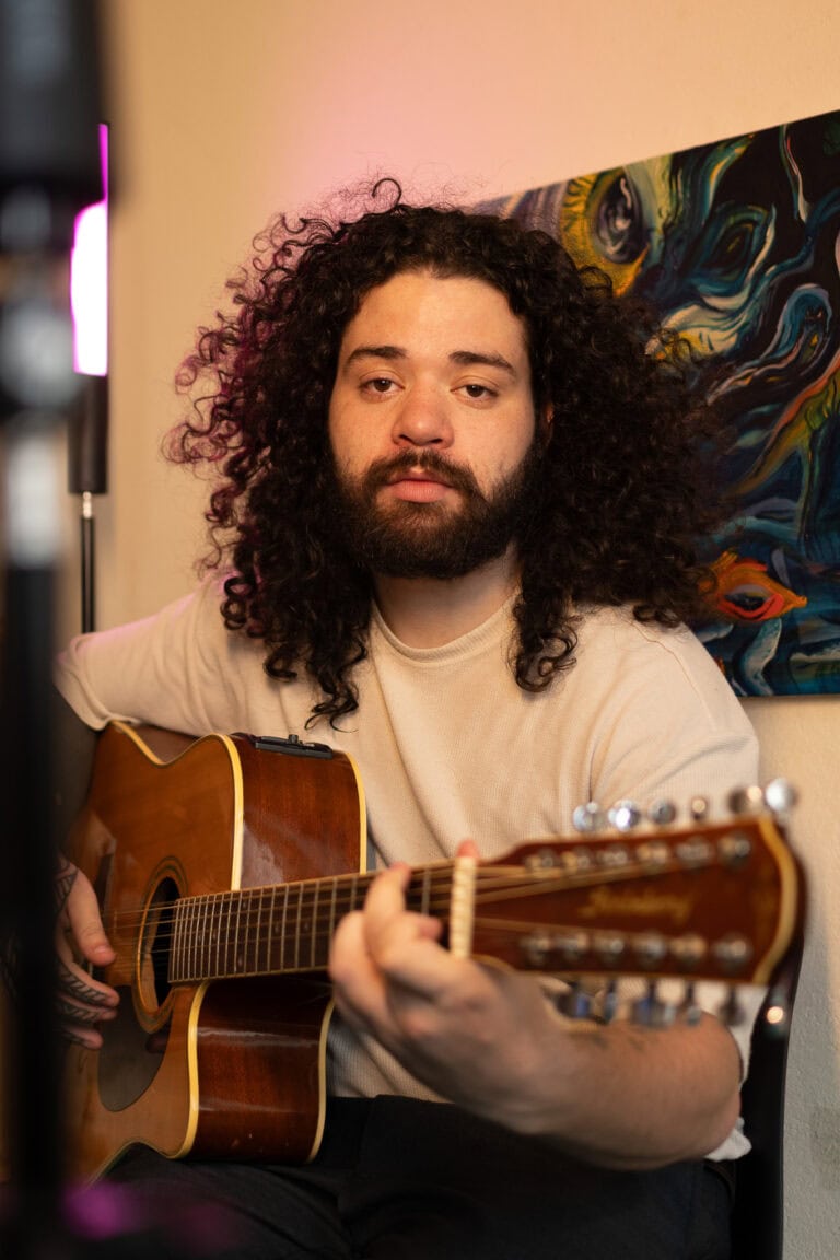 Imagem de um homem com cabelo cacheado tocando violão em ambiente interno.