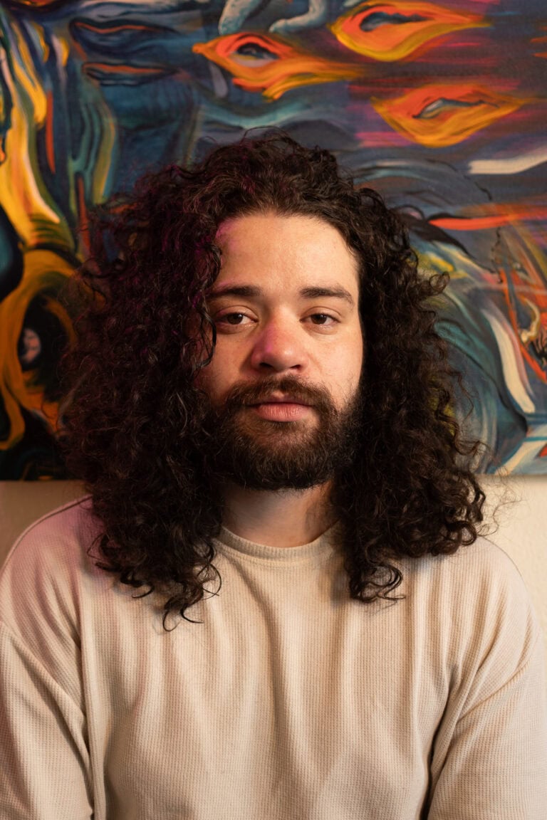 Imagem de retrato de um homem com cabelo cacheado e barba, em ambiente com arte abstrata ao fundo.