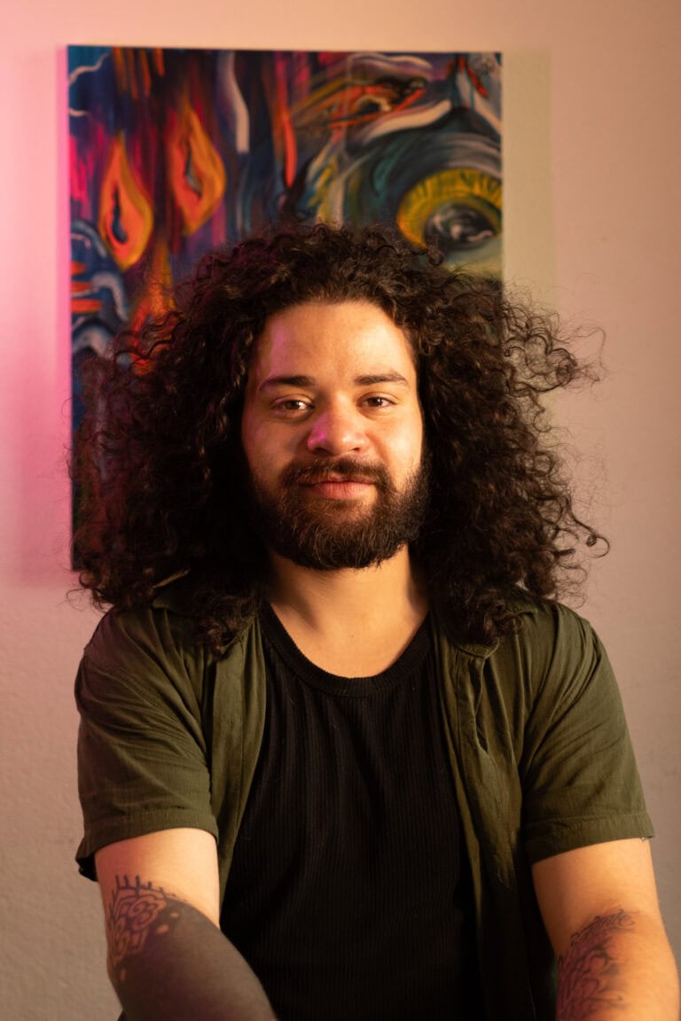 Fotógrafo Guilherme Marques com cabelo cacheado e barba, posando em estúdio com obra de arte ao fund.