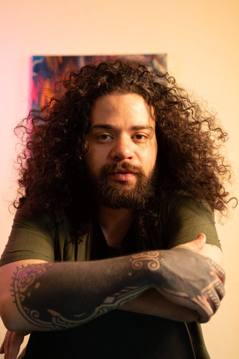 Fotógrafo Guilherme Marques com cabelo cacheado e tatuagens, retrato profissional.