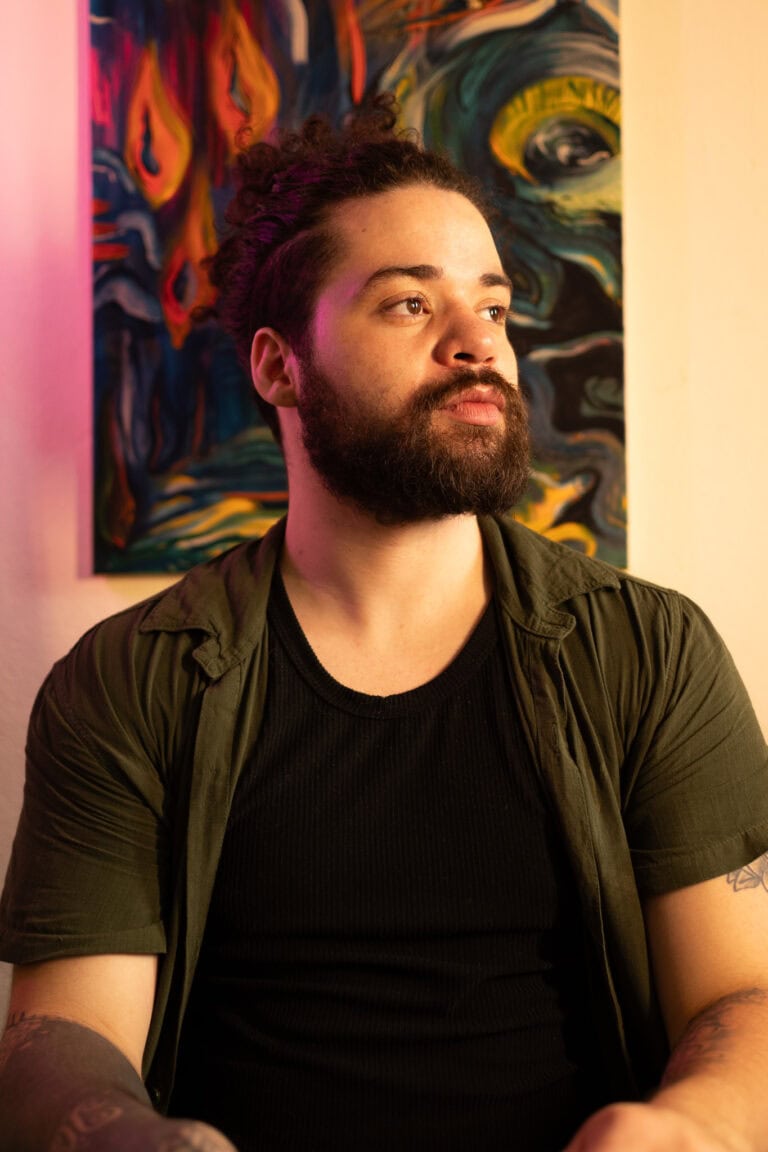 Retrato de homem com barba e cabelo cacheado, em ambiente artístico.