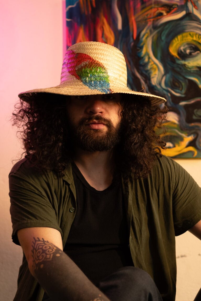 Retrato de fotógrafo com chapéu de palha, cabelo cacheado e tatuagem no braço, em ambiente artístico.