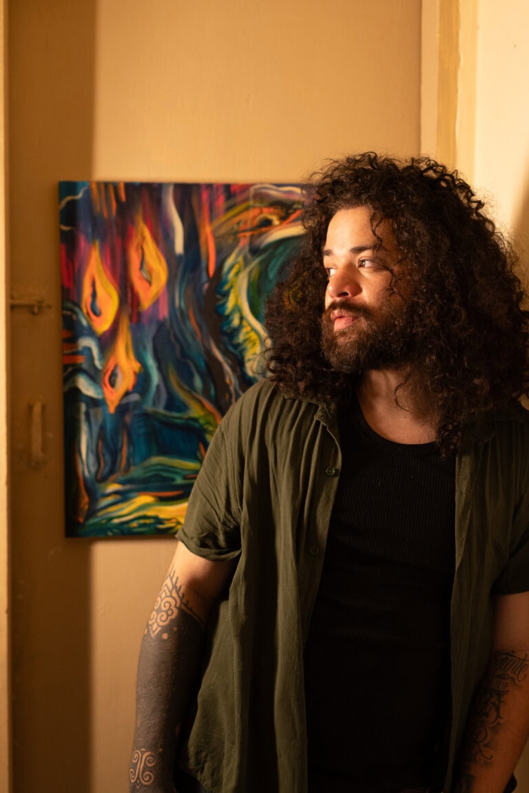 Imagem de um homem com cabelo cacheado e barba, posando ao lado de uma obra de arte colorida.