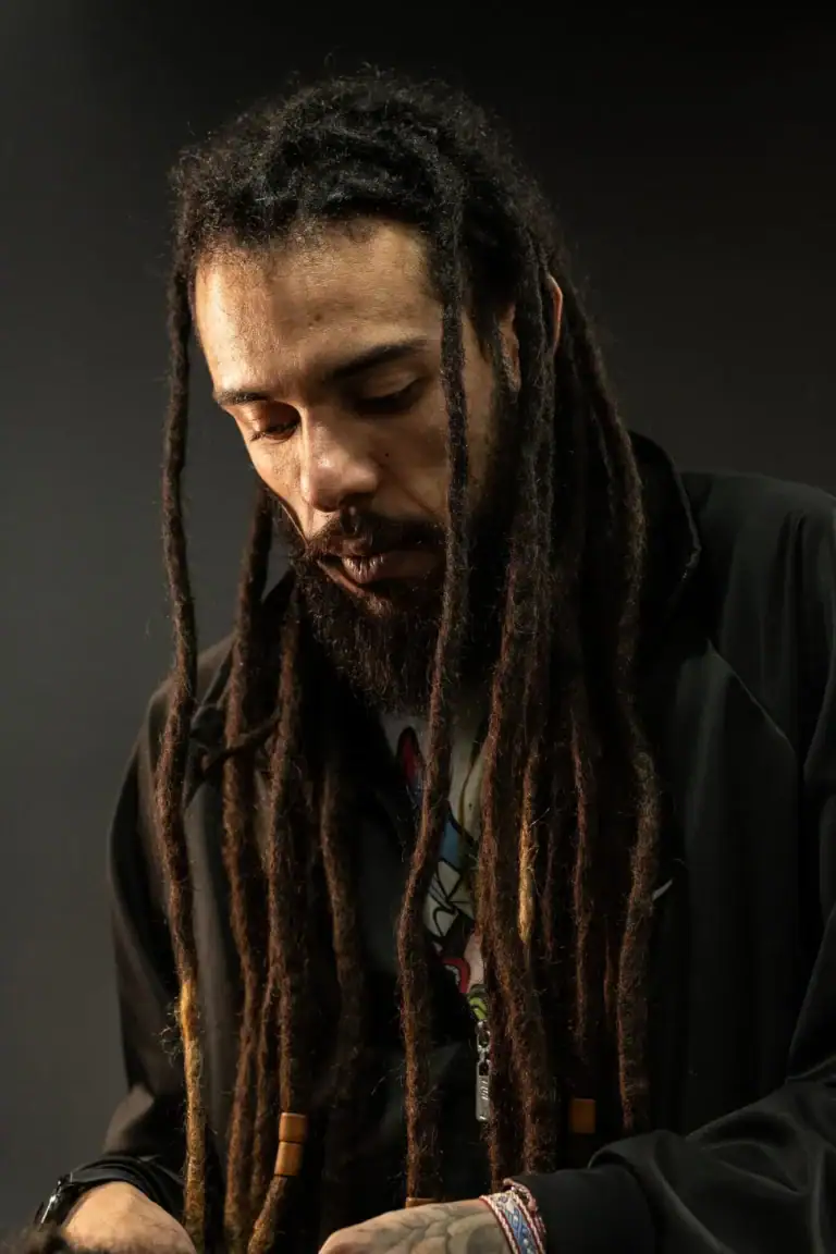 Retrato de um homem com dreadlocks e expressão pensativa, fundo escuro.