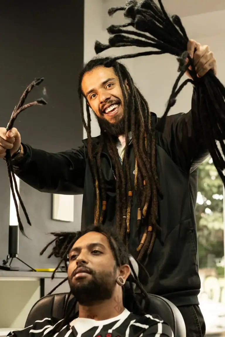 Fotógrafo com dreadlocks sorrindo, segurando cabelo, em estúdio com fundo neutro.