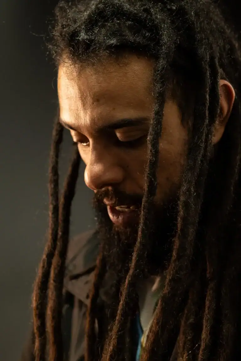 Retrato de um homem com dreadlocks, expressão pensativa, fundo escuro.