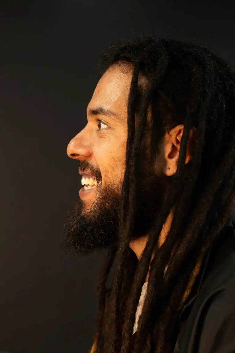 Retrato de um homem com dreadlocks sorrindo, perfil, fundo escuro.