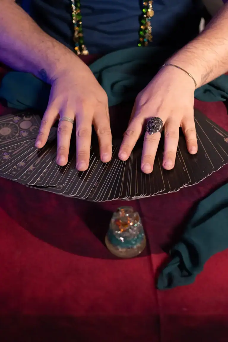 Mãos com anéis e cartas de tarô sobre mesa vermelha.
