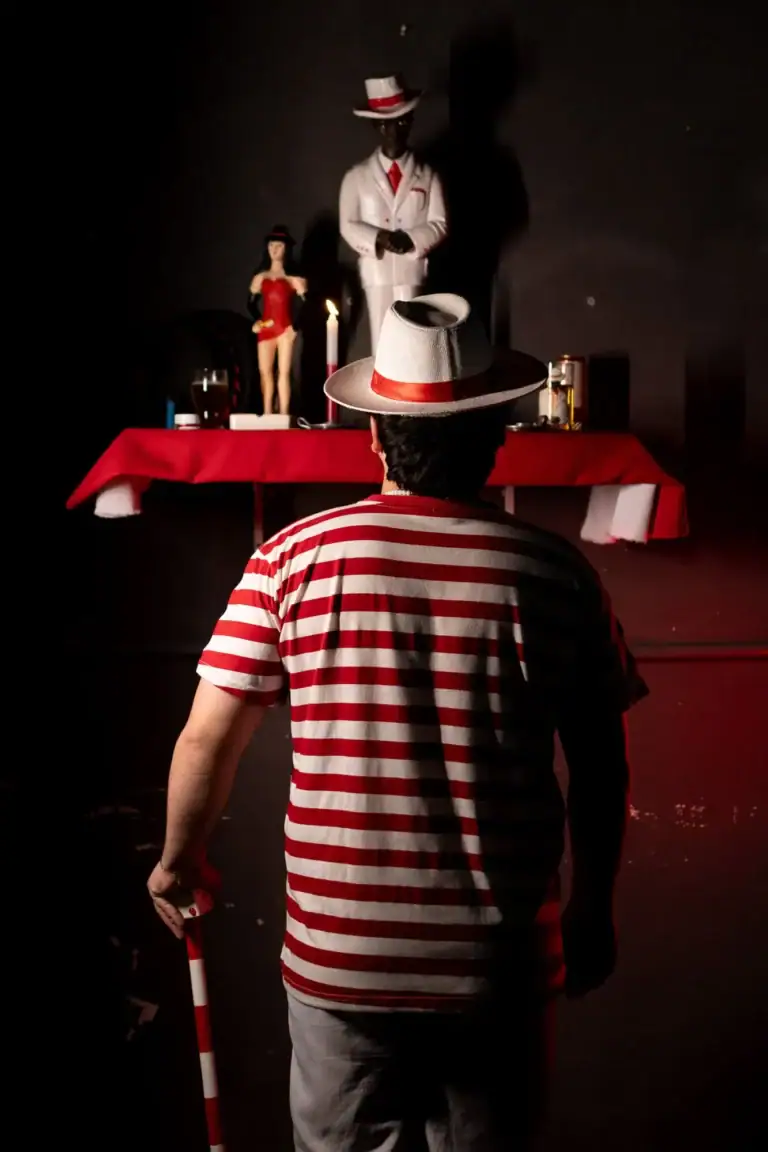Homem com camiseta listrada vermelha e branca diante de altar com figuras e objetos.