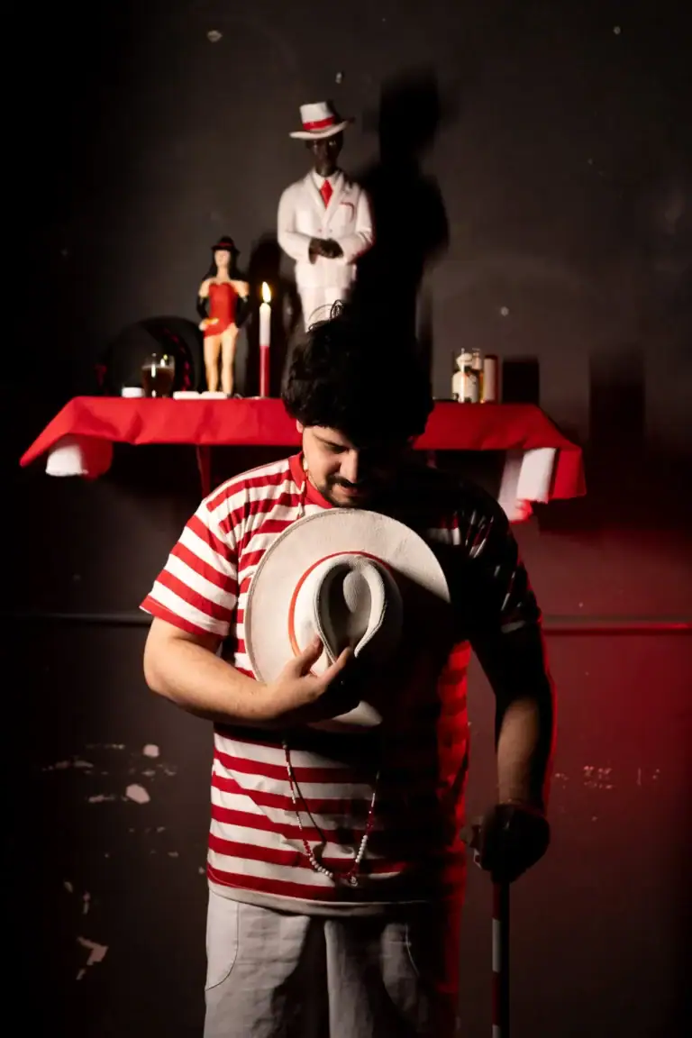 Homem com chapéu e bastão, cenário teatral com figuras e velas ao fundo.
