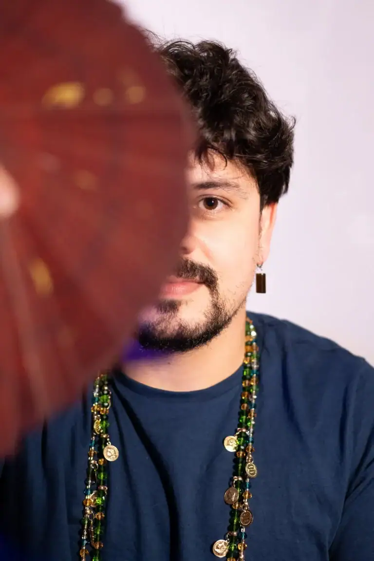 Retrato de um homem com cabelo escuro e barba, usando colar de moedas, com fundo branco.