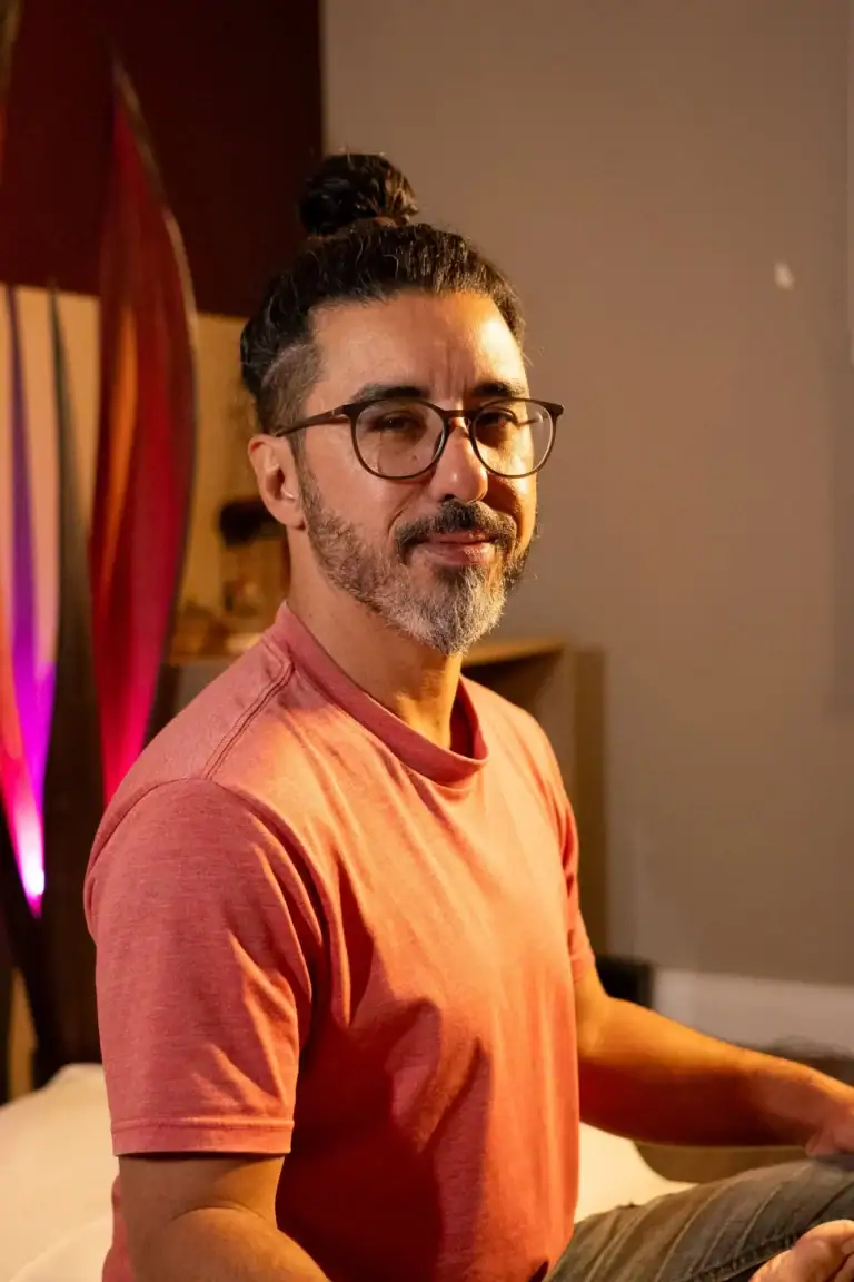 Retrato de um homem com cabelo preso, barba e óculos, usando camiseta laranja.